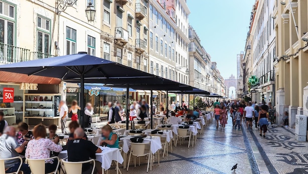 A bustling outdoor café with tables and chairs, set against a backdrop of historic buildings and a cobblestone street.