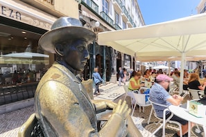 Une statue de bronze représentant un homme portant un chapeau, située dans un espace extérieur d’un café, avec des tables et des parasols.