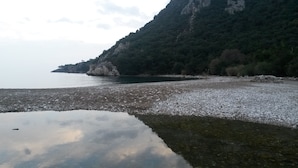 A natural landscape with a rocky shoreline, calm water, and a forested hill in the background. - Hotel Yavuz Bungalow (Kemer)