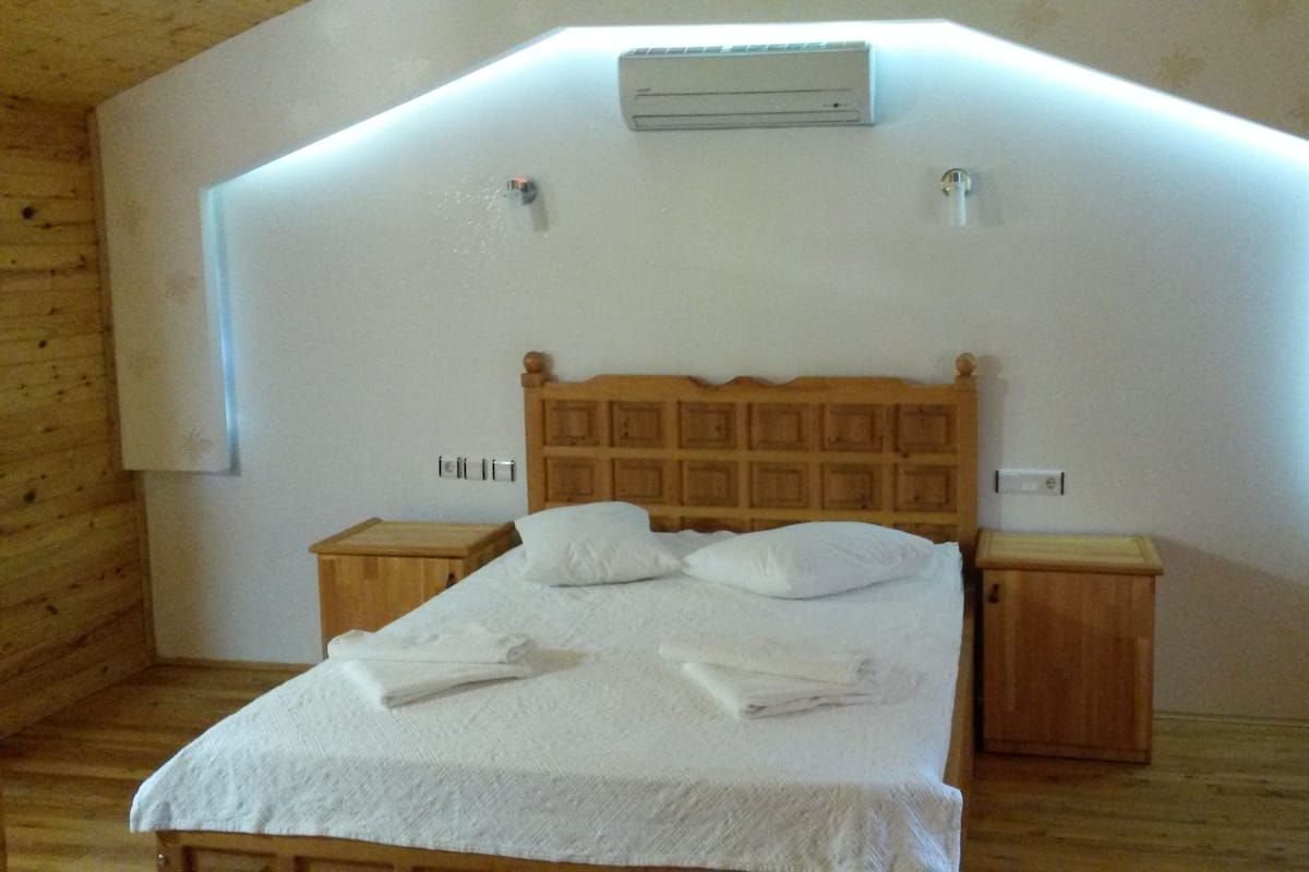 Quarto com cama de madeira, roupa de cama branca, duas mesas de cabeceira e teto inclinado com unidade de ar condicionado.