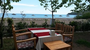 A table set with a red and white tablecloth, wooden chairs with patterned cushions, and a view of the beach with umbrellas and rocks. - Hotel Yavuz Bungalow (Kemer)