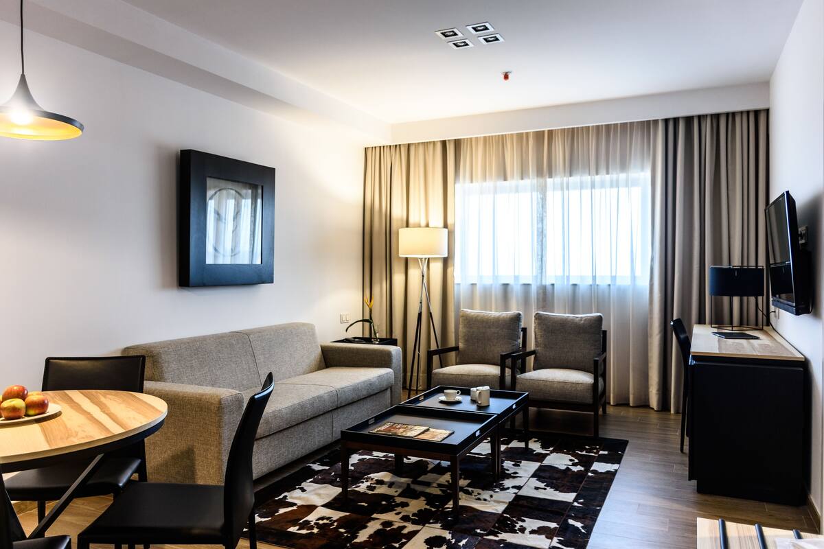 Habitación de hotel moderna con sofá, dos sillones, una mesa de comedor con manzanas, una mesita de centro, una lámpara, un televisor y un amplio ventanal con cortinas.