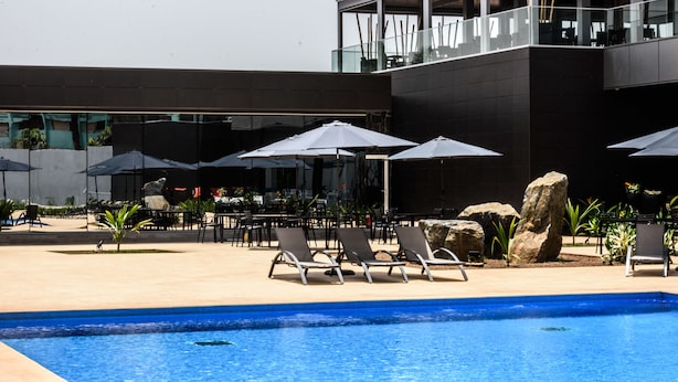 Un espace piscine moderne avec des chaises longues et des parasols, une piscine à l’eau d’un bleu limpide et un bâtiment doté de grandes fenêtres et d’un balcon.