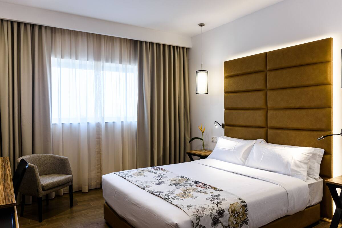 Una habitación de hotel con una cama, una silla, una mesita de noche, una lámpara y un ventanal con cortinas.