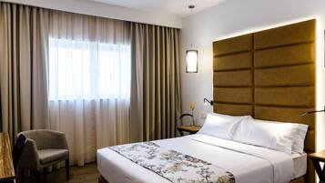 Una habitación de hotel con una cama, una silla, una mesita de noche, una lámpara y un ventanal con cortinas.