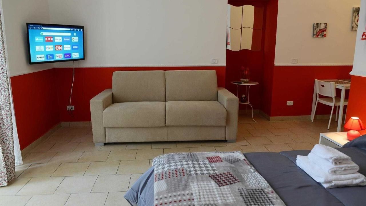 A room with a sofa, a bed with a blanket, a TV mounted on the wall, and a small table with a lamp.