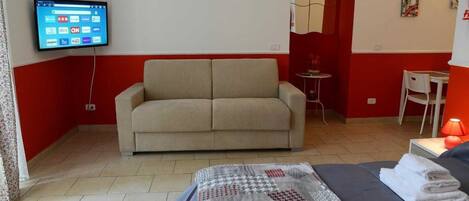 A room with a sofa, a bed with a blanket, a TV mounted on the wall, and a small table with a lamp.