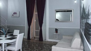 A living room with a white sofa, a dining table with chairs, a small side table, a mirror, and a patterned carpet. - B&B La Torretta di Chiaia (Naples)