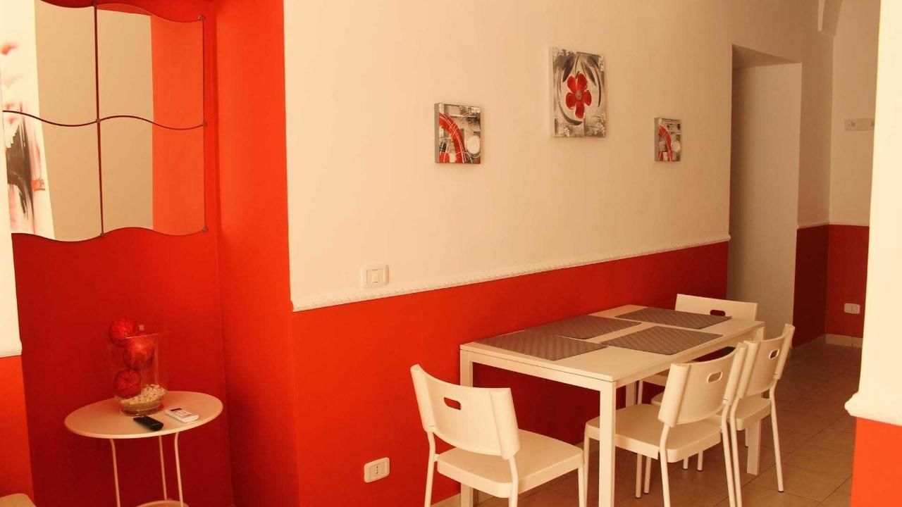 A dining area with a white table and chairs, a small side table with a vase and remote control, and a red and white wall with three framed pictures.