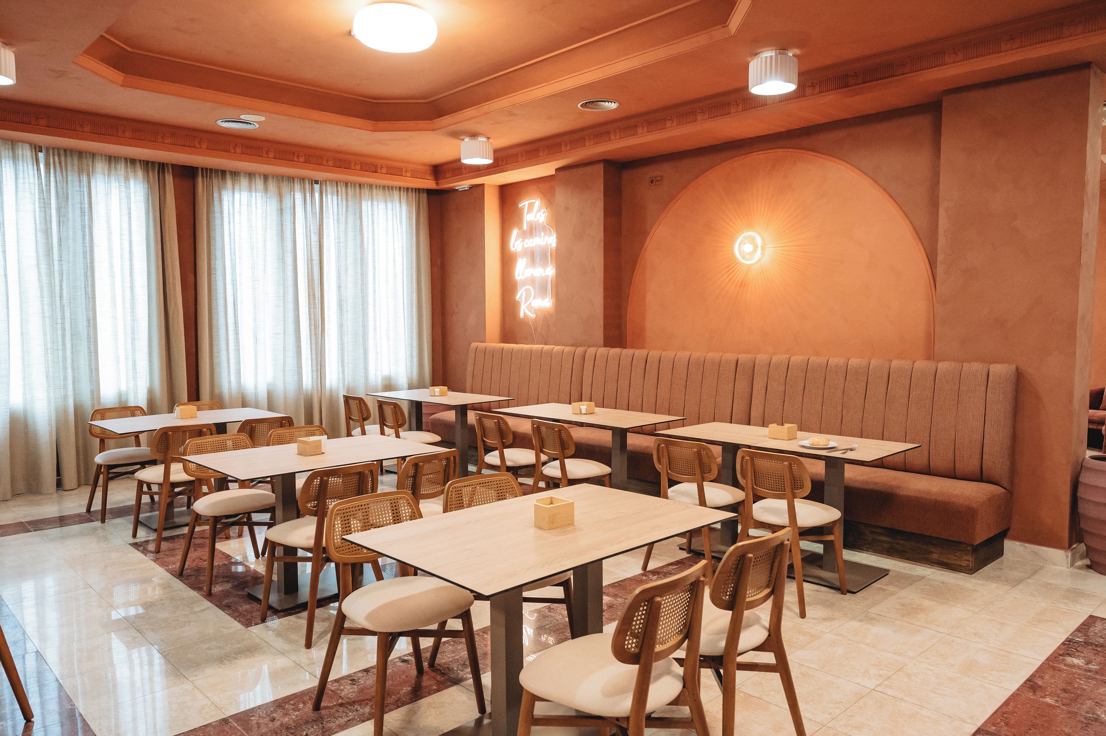 Un comedor de hotel con varias mesas y sillas, un letrero de neón en la pared y un amplio ventanal de arco.