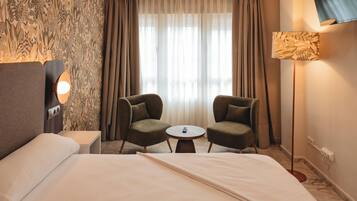 A hotel room with a bed, two armchairs, a small table, a TV, and a window with curtains.