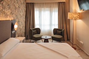 A hotel room with a bed, two armchairs, a small table, a TV, and a window with curtains. - Roma Aurea (Talavera de la Reina)