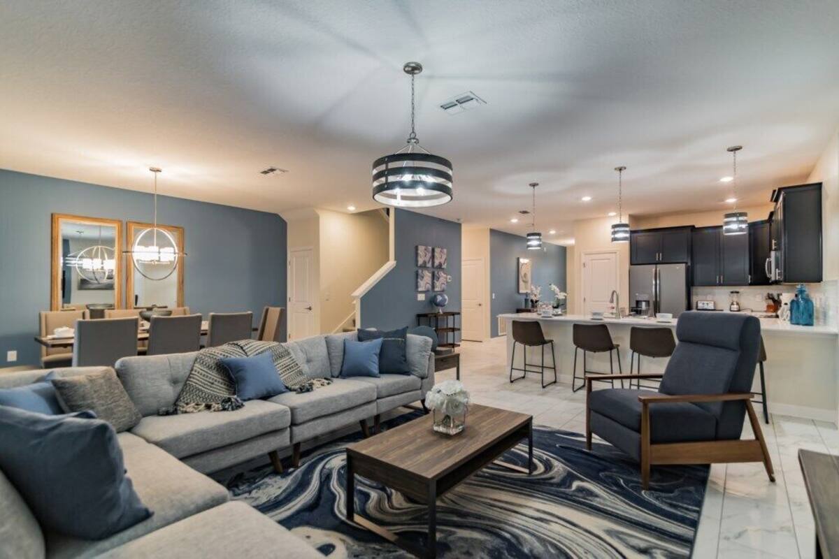 A modern living room with a sofa, coffee table, and a dining area with chairs. There is a kitchen with bar stools and a refrigerator.
