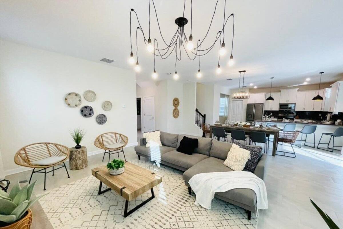 A spacious living room with a sofa, coffee table, and wicker chairs. A kitchen is visible in the background with modern appliances and pendant lighting.