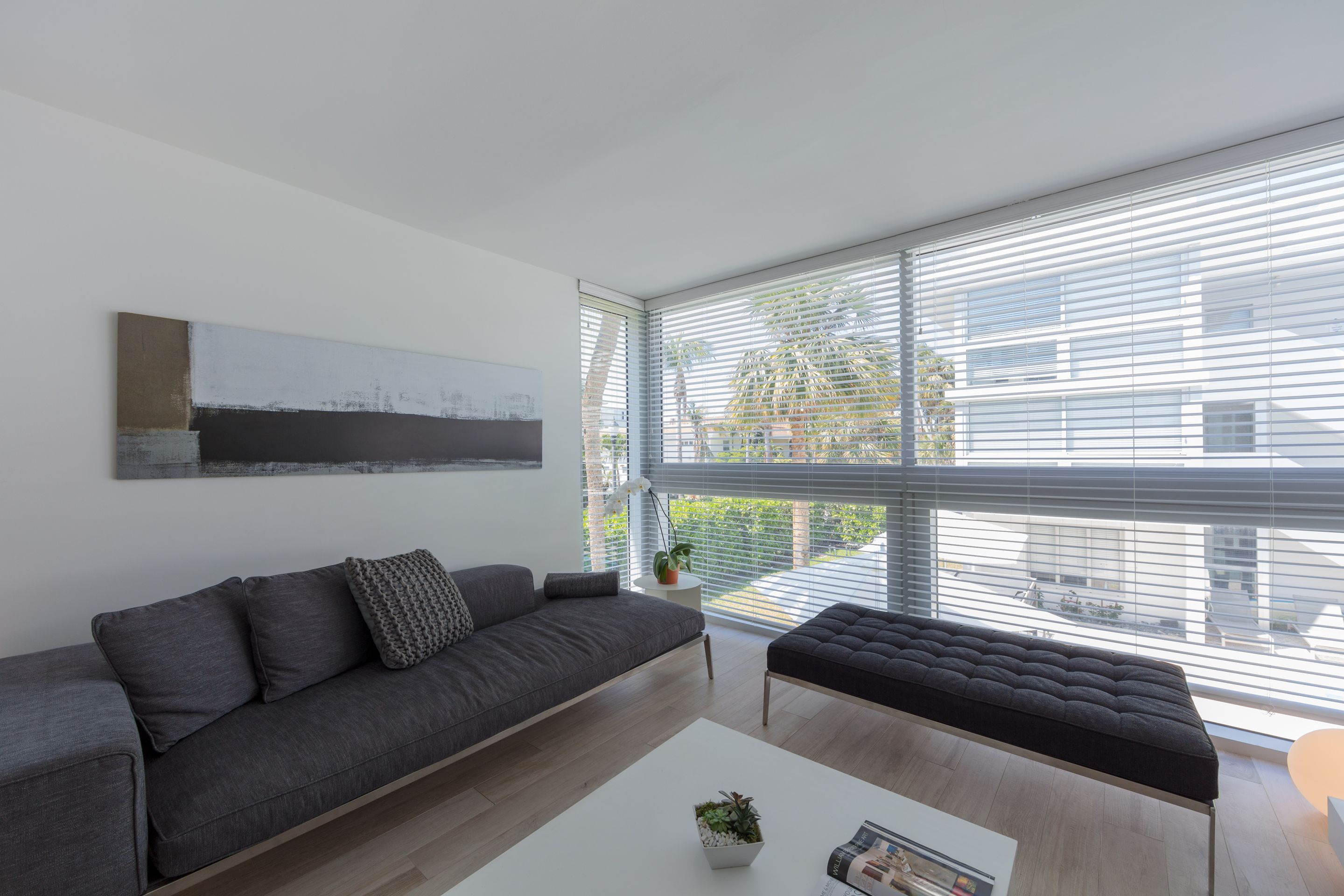 A modern living room with a grey sofa, a black ottoman, a white coffee table with a plant, and a large window with blinds.