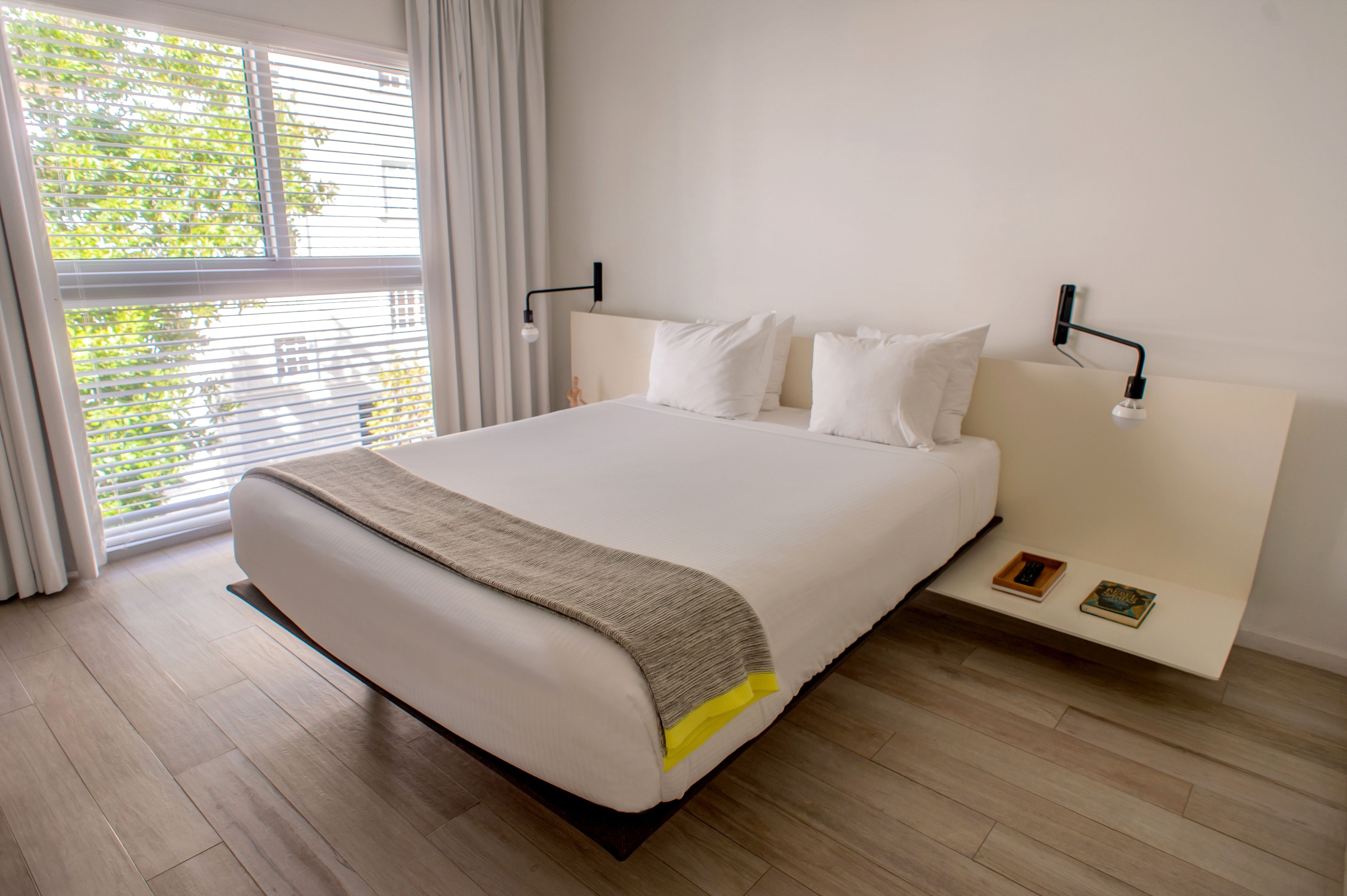 A bedroom with a large bed, white bedding, a bedside table with books, and a window with blinds.