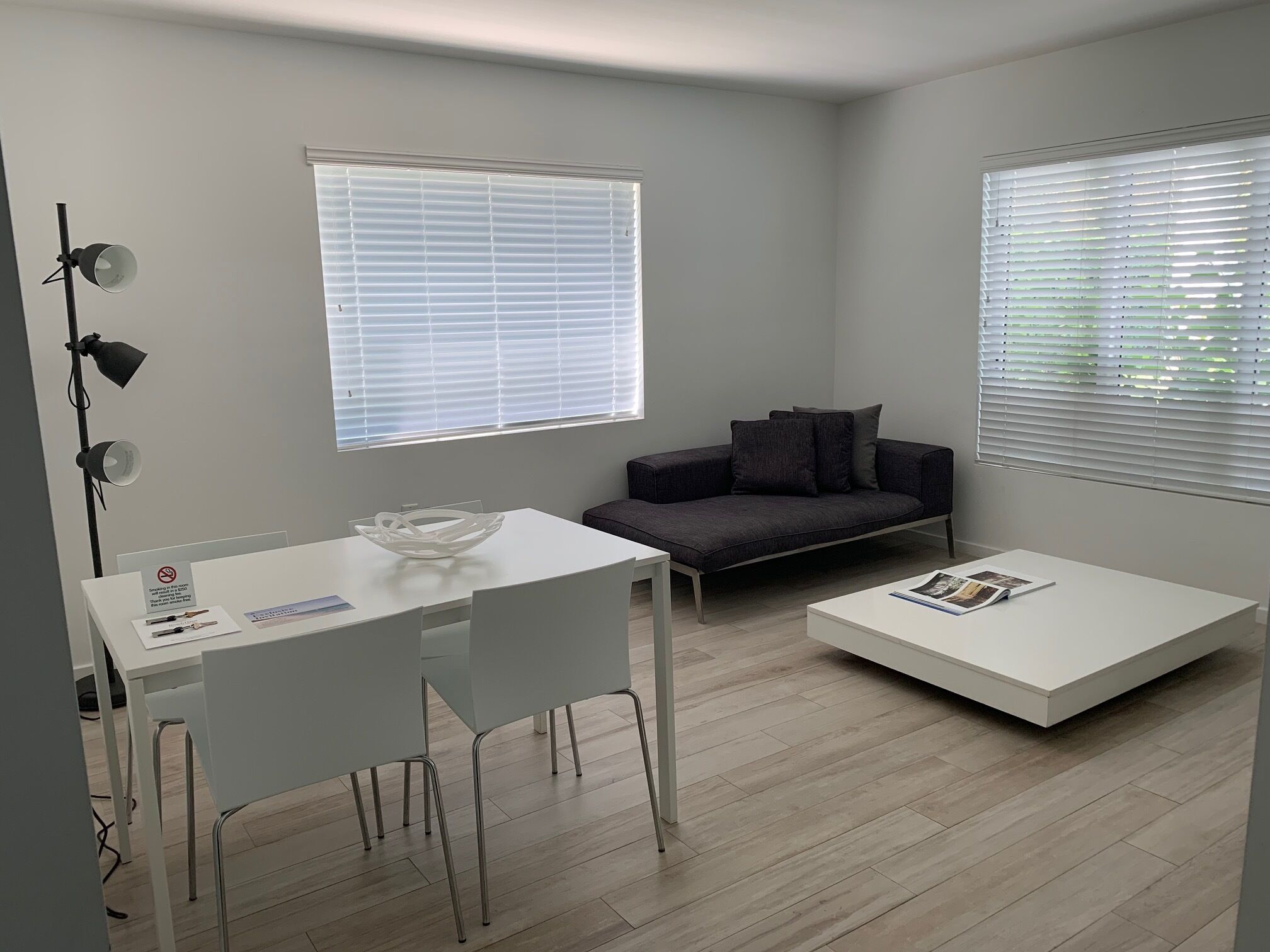 A modern living room with a white dining table, a dark grey sofa, and a white coffee table. There are two windows with blinds, a floor lamp, and a sign on the table.