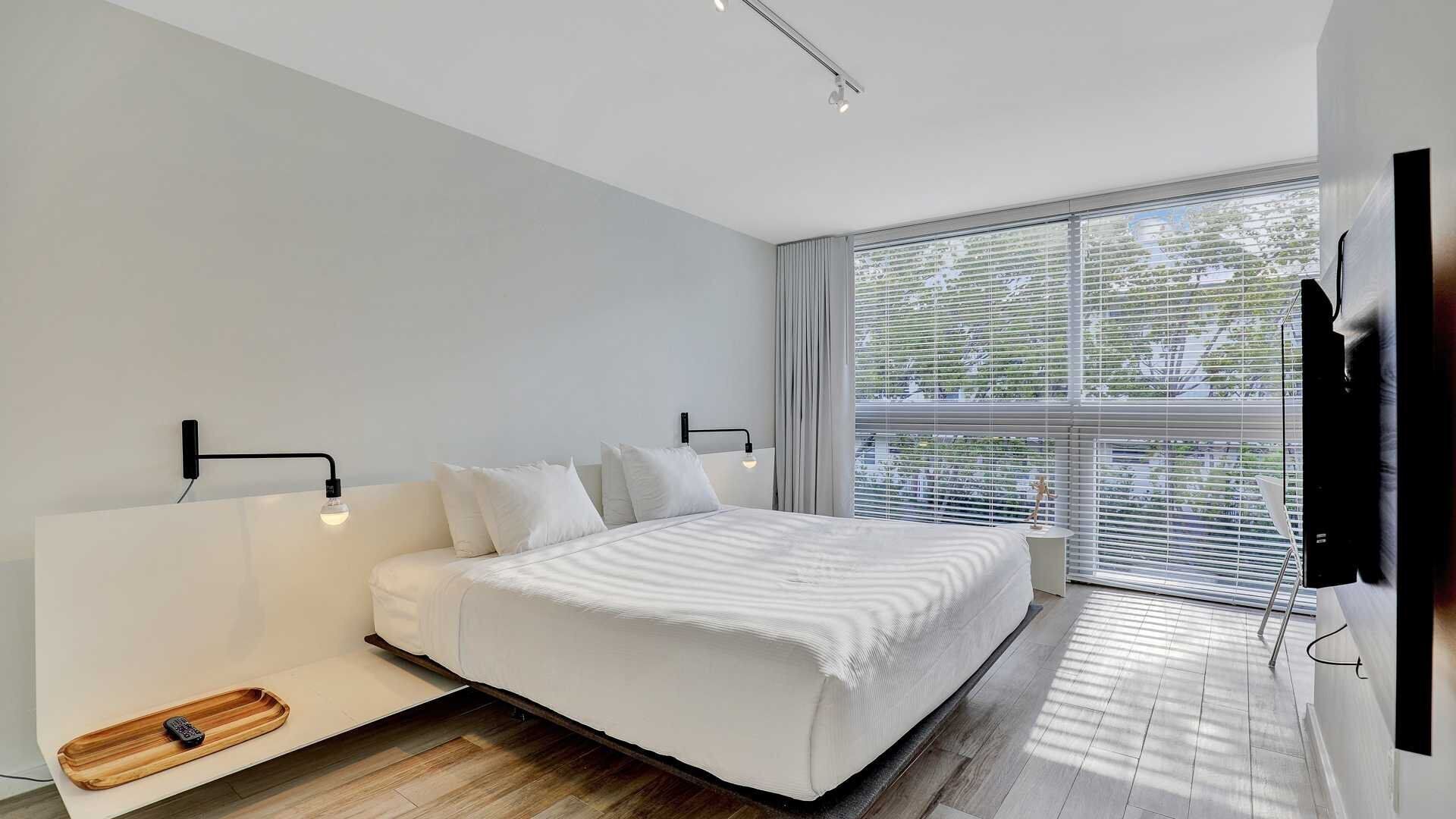 Un dormitorio moderno con una cama grande, una mesita de noche, un televisor y una ventana con persianas.