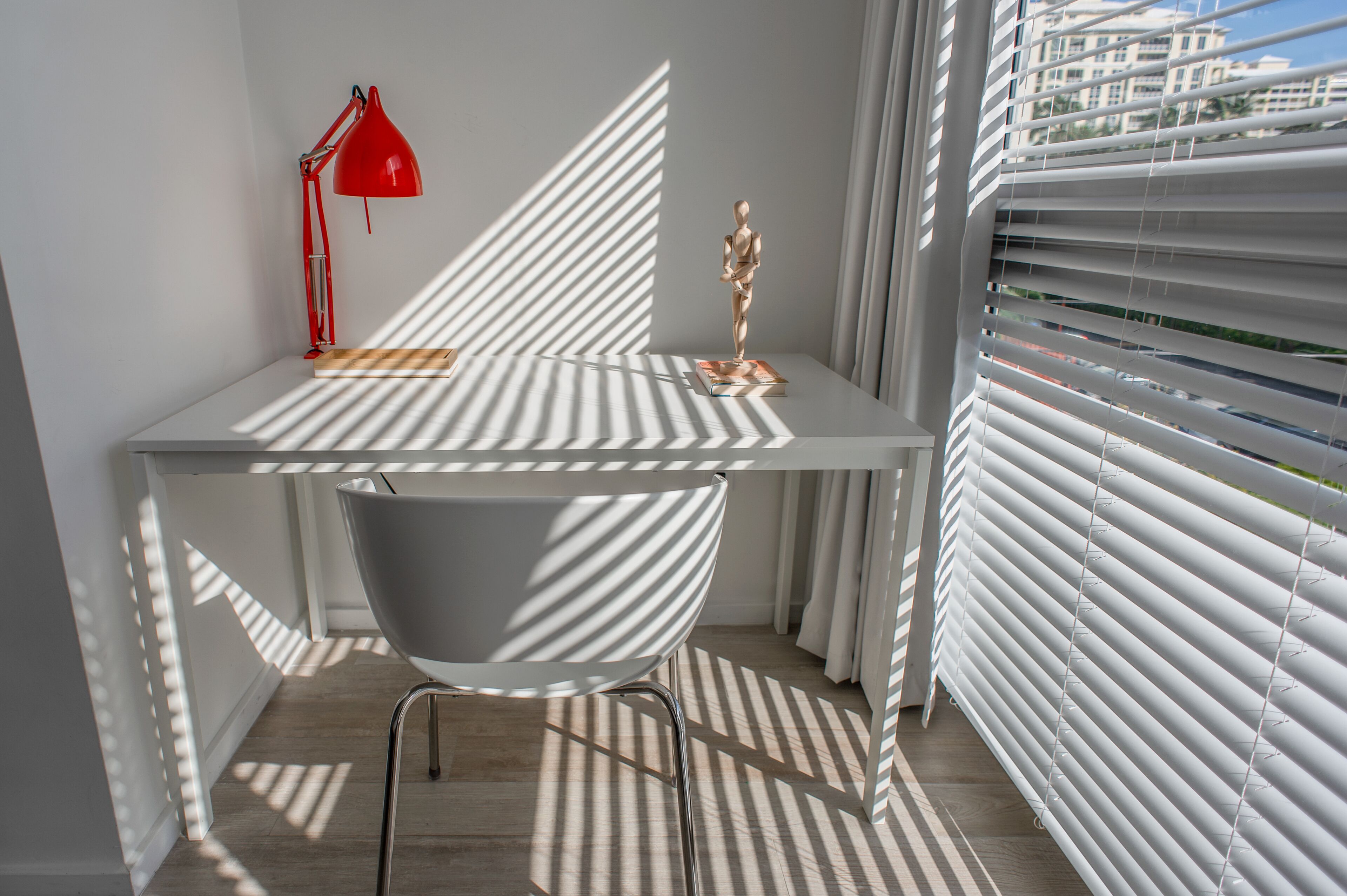 A modern room with a white desk, a red desk lamp, a white chair, and a window with blinds.