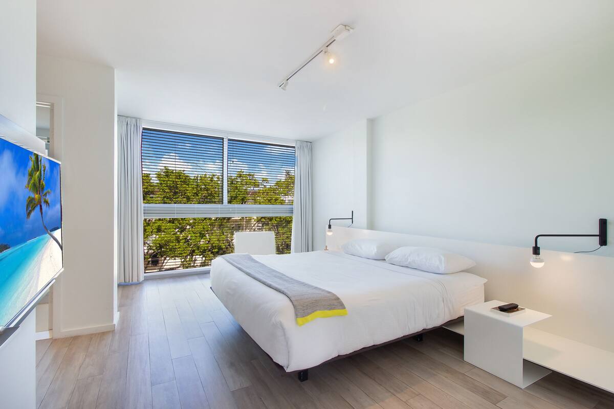 A modern bedroom with a large bed, a bedside table, a TV, and a window with blinds.