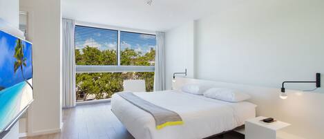 A modern bedroom with a large bed, a bedside table, a TV, and a window with blinds.