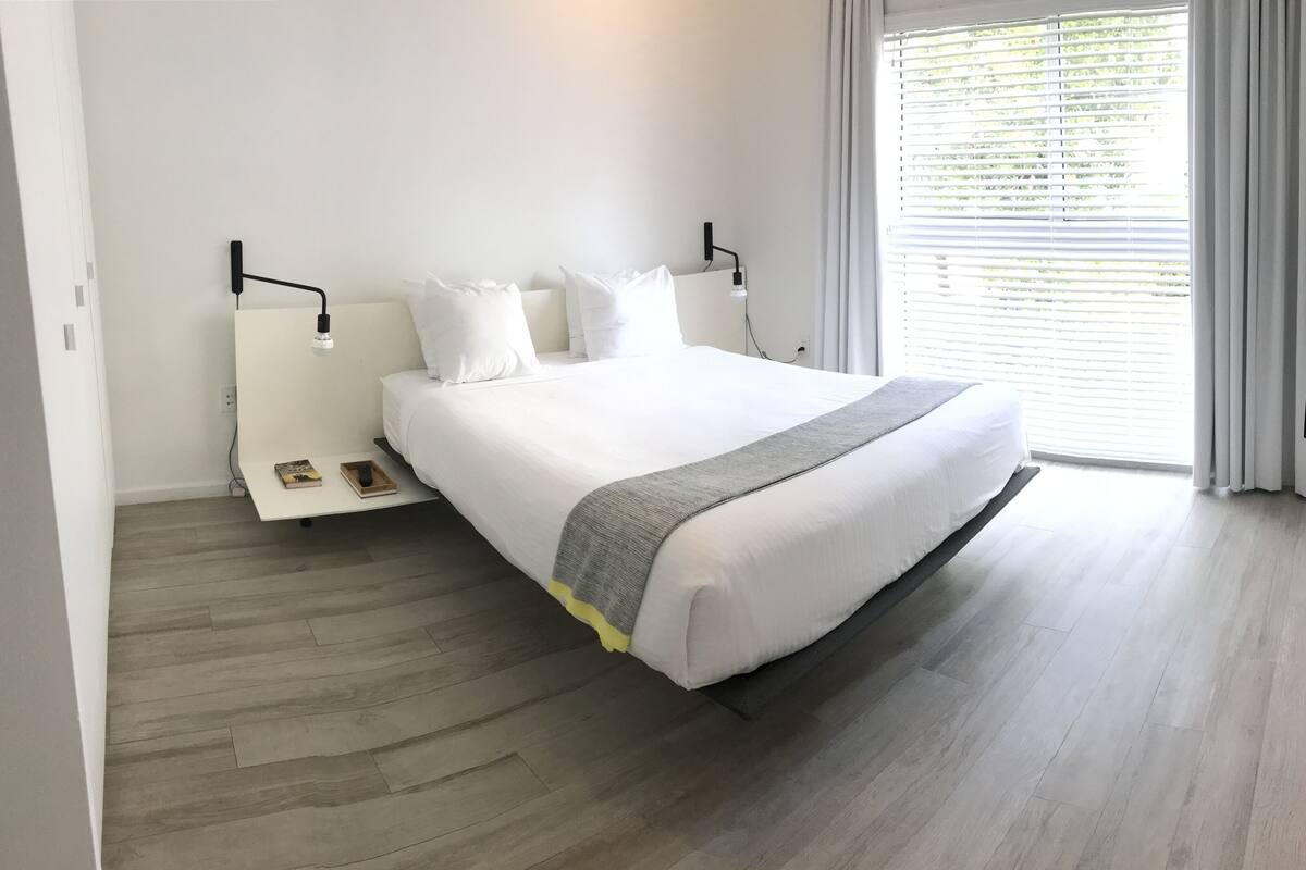 A modern hotel room with a large bed, a bedside table with a book and remote, a wall-mounted TV, and a window with blinds.