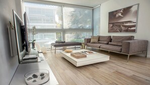 A modern living room with a flat-screen TV, a sofa, a coffee table, and a large window with blinds. - Beach Haus Key Biscayne (Key Biscayne)