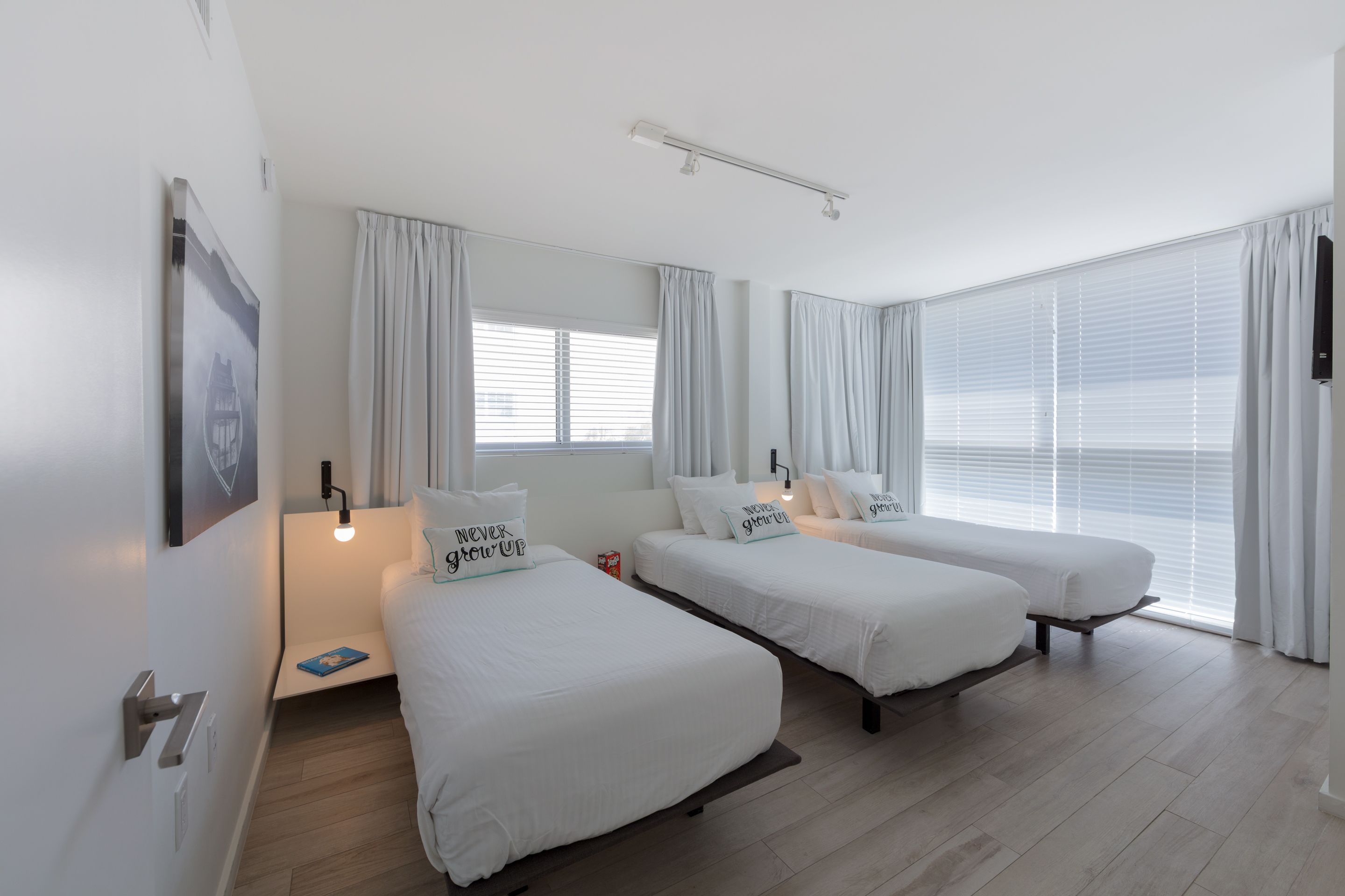 A hotel room with two single beds, a bedside table with a lamp, a large window with curtains, and a wall-mounted TV.