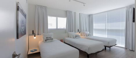 A hotel room with two single beds, a bedside table with a lamp, a large window with curtains, and a wall-mounted TV.