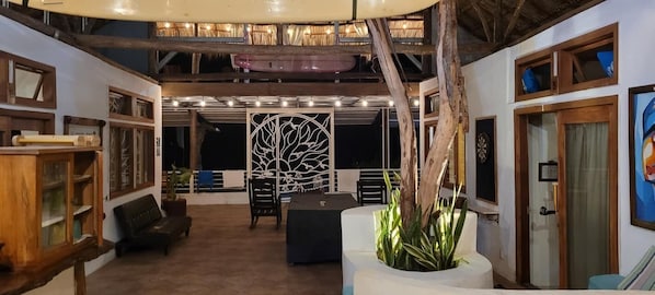 An indoor space with a high ceiling, wooden beams, and a central decorative panel. There are seating areas with cushions and a potted plant.