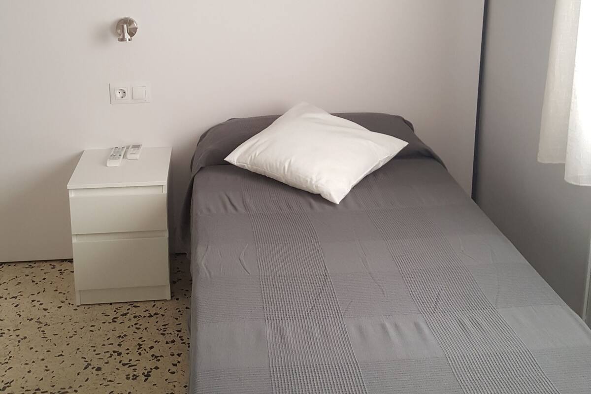 Une petite chambre avec un lit simple, une couvre-lit grise, un oreiller blanc, une table de chevet avec une télécommande et un climatiseur fixé au mur.