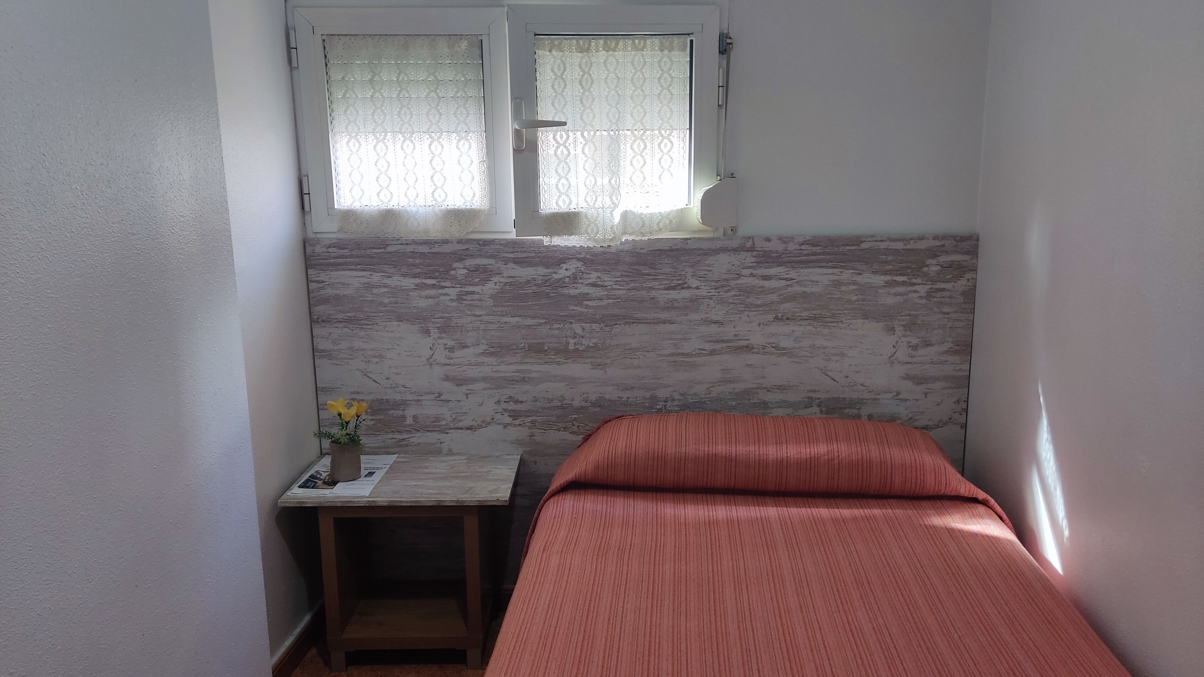 Una habitación pequeña con una cama, una mesita de noche de madera con un jarrón de flores y una ventana con cortinas de encaje.