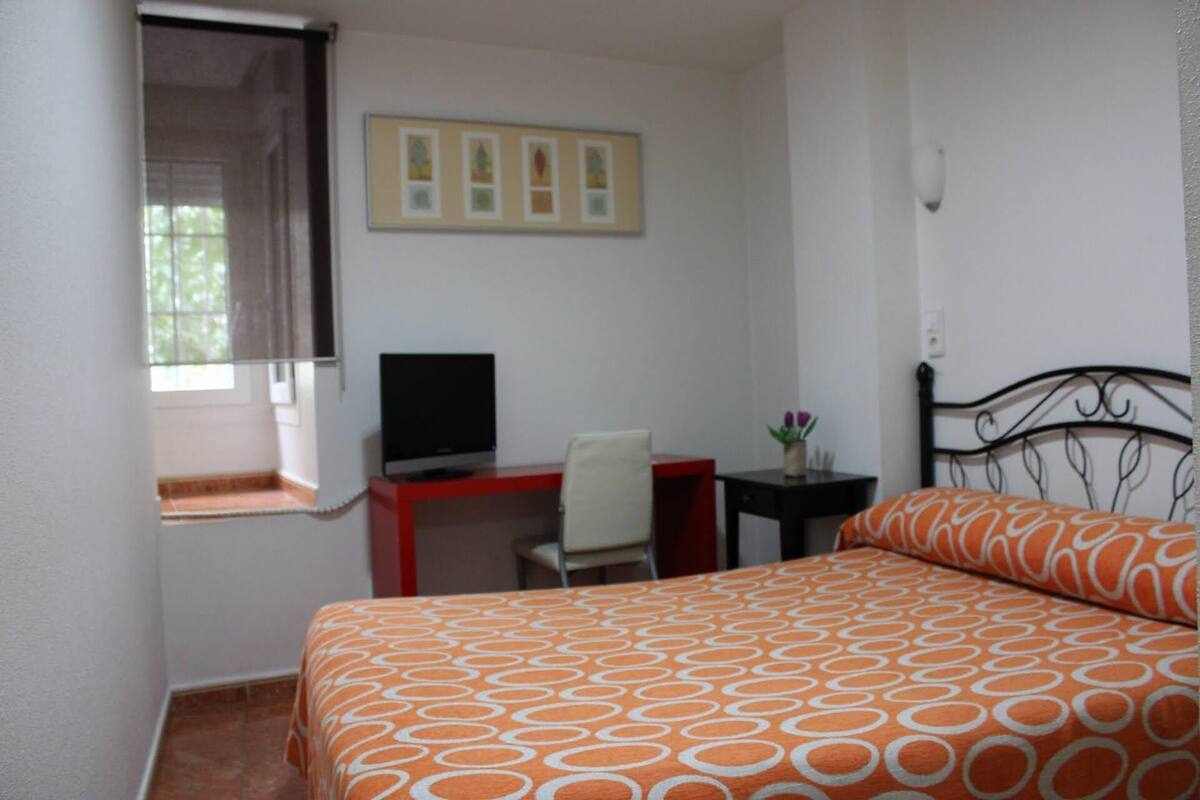 A bedroom with a bed, a desk, a chair, a TV, and a window.