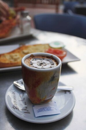 Una taza de café sobre un plato con una bandeja de comida al fondo.