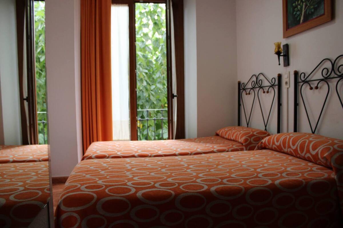 A hotel room with two beds, a window with curtains, a metal headboard, and a wall-mounted light fixture.
