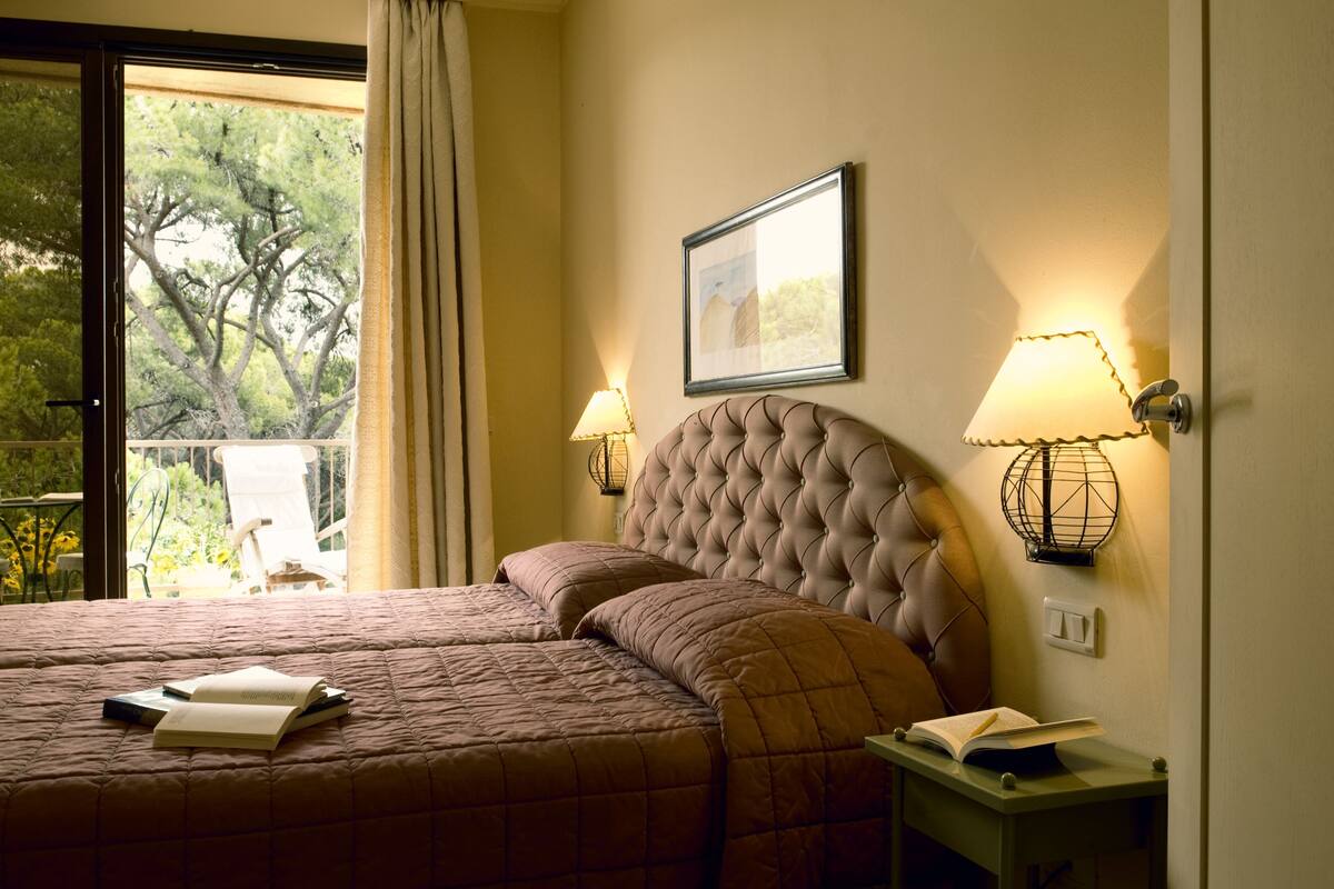 A bedroom with a large bed, two bedside lamps, a framed picture on the wall, and a view of trees through a window.