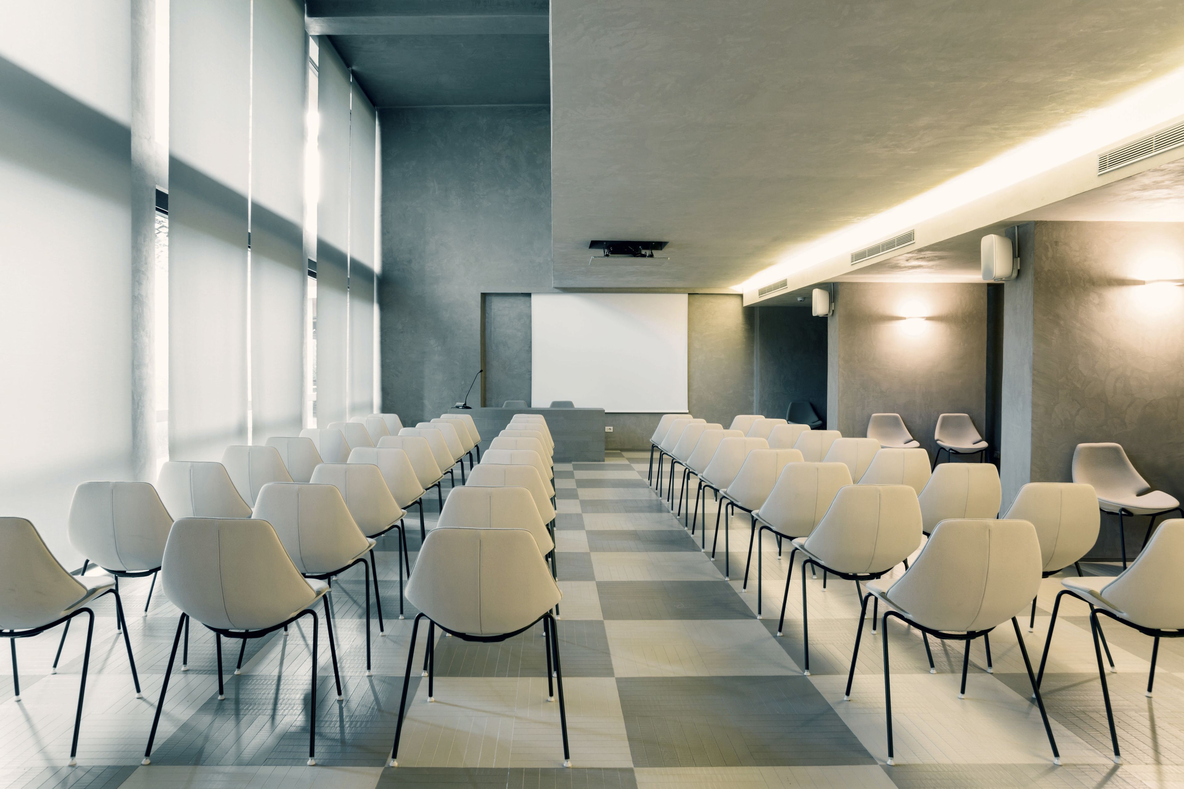 Una sala conferenze con file di sedie rivolte verso uno schermo di proiezione, ampie finestre e un pavimento a scacchi.