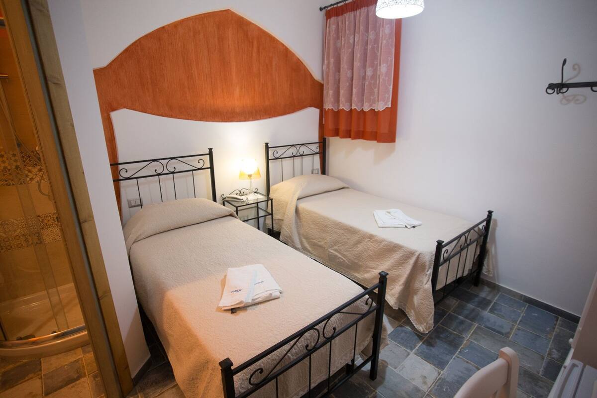 Una habitación con dos camas individuales, una mesita de noche con lámpara, zona de ducha y ventana con cortina.