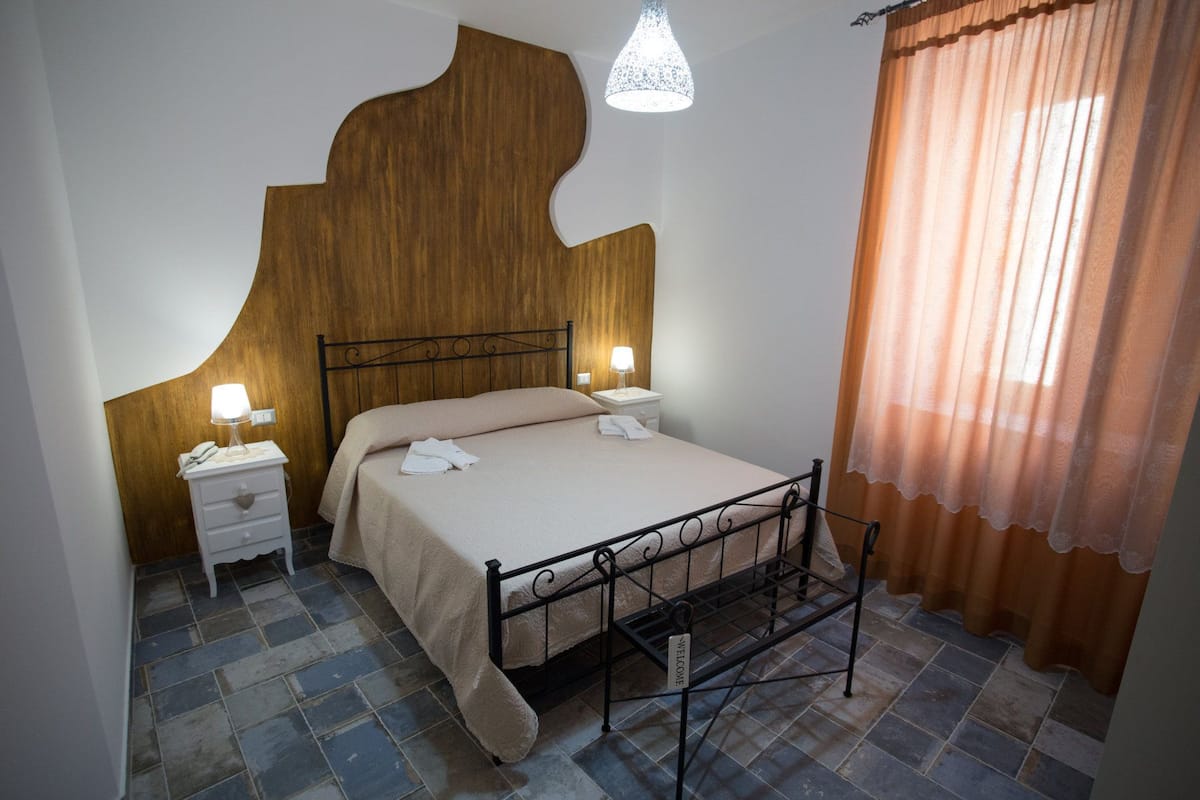 Un dormitorio con una cama, dos mesitas de noche con lámparas, un estante junto a la cama y una cortina.