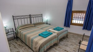 Una cama doble con ropa de cama a rayas y dos toallas enrolladas, una mesita de noche con lámpara, y una ventana con cortinas azules.