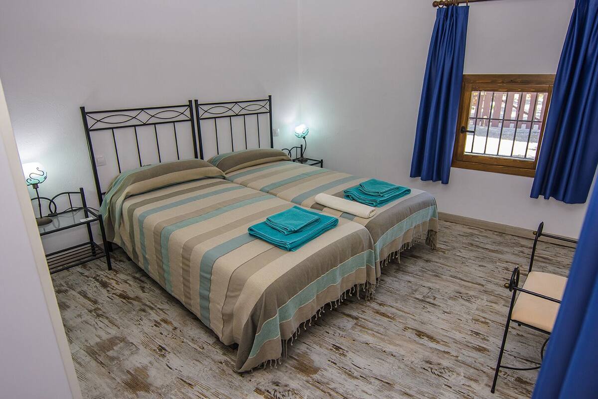 A double bed with striped bedding and two rolled towels, a bedside table with a lamp, and a window with blue curtains.