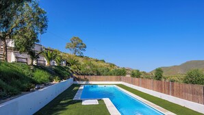 Una piscina rectangular con borde blanco, rodeada de césped artificial, ubicada en una colina bajo un cielo azul despejado.