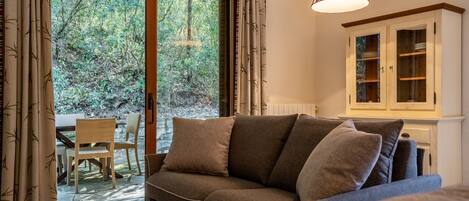 A cozy living area with a sofa, a dining table with chairs, and a view of a forest through a glass door.