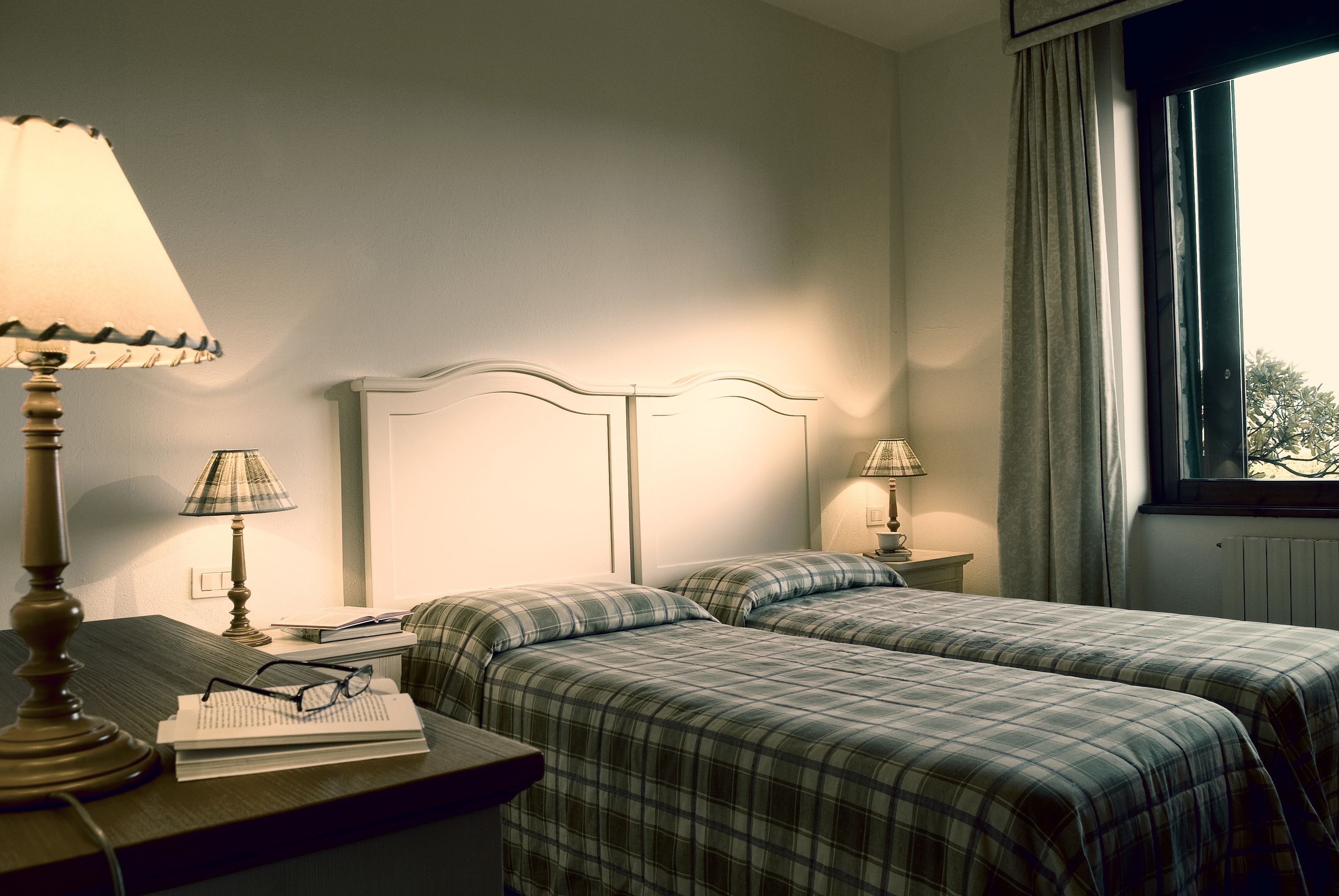 A hotel room with two beds, a nightstand with a lamp, a window with curtains, and a view of trees outside.