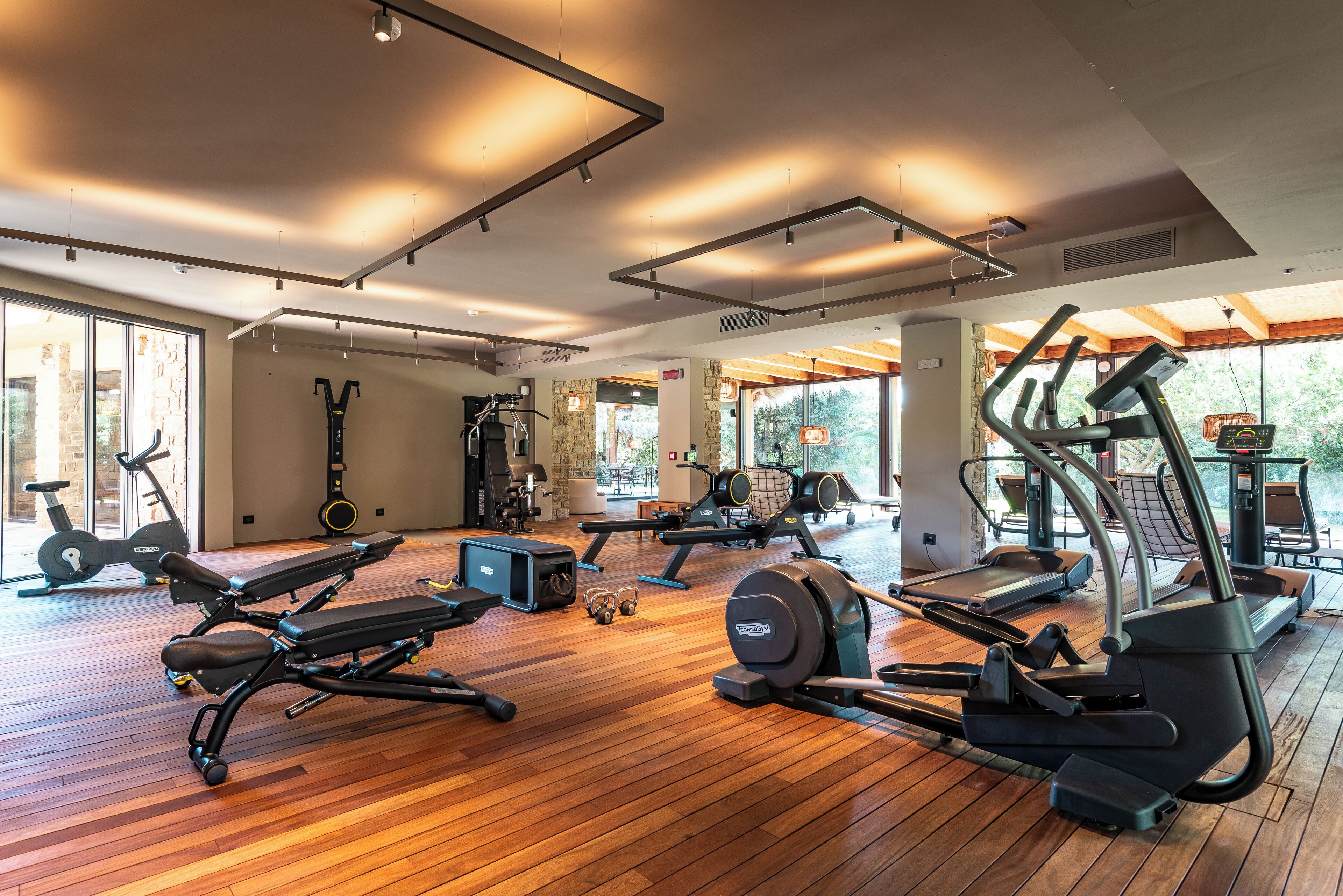 Een sportschool met diverse trainingsapparaten, grote ramen en een houten vloer.