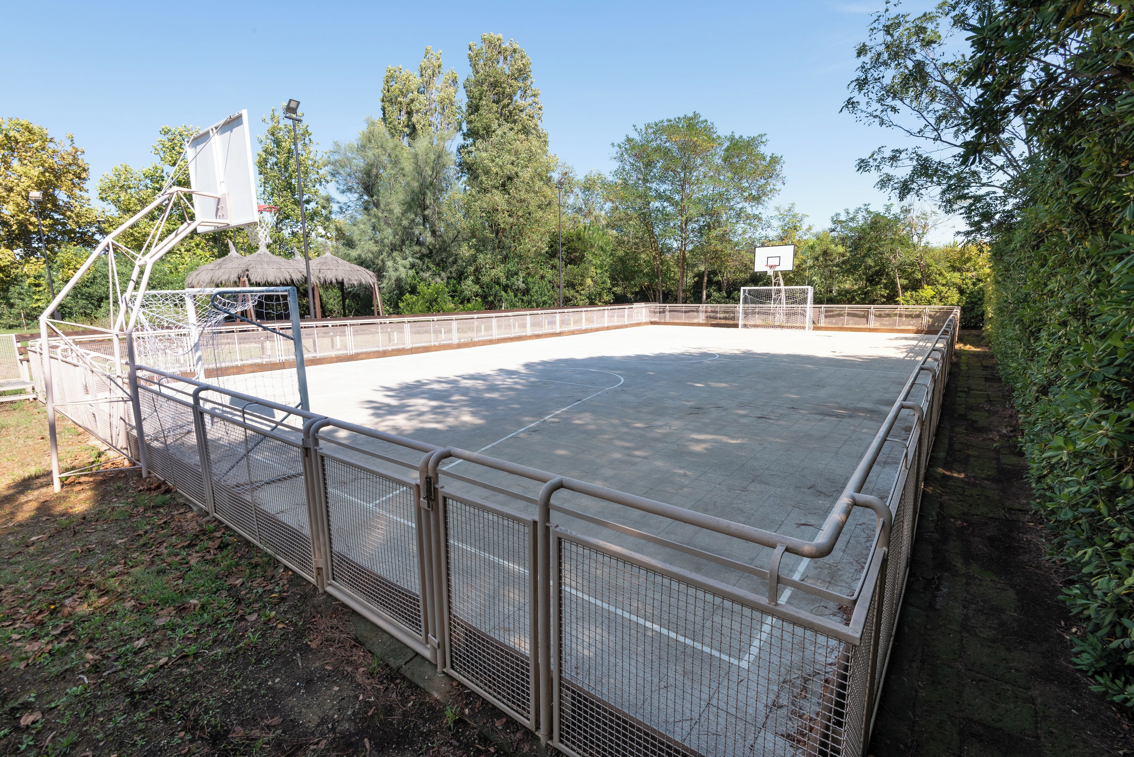 Un terrain de basket en plein air, entouré d’une clôture en métal, avec un panier de basket et un petit abri.