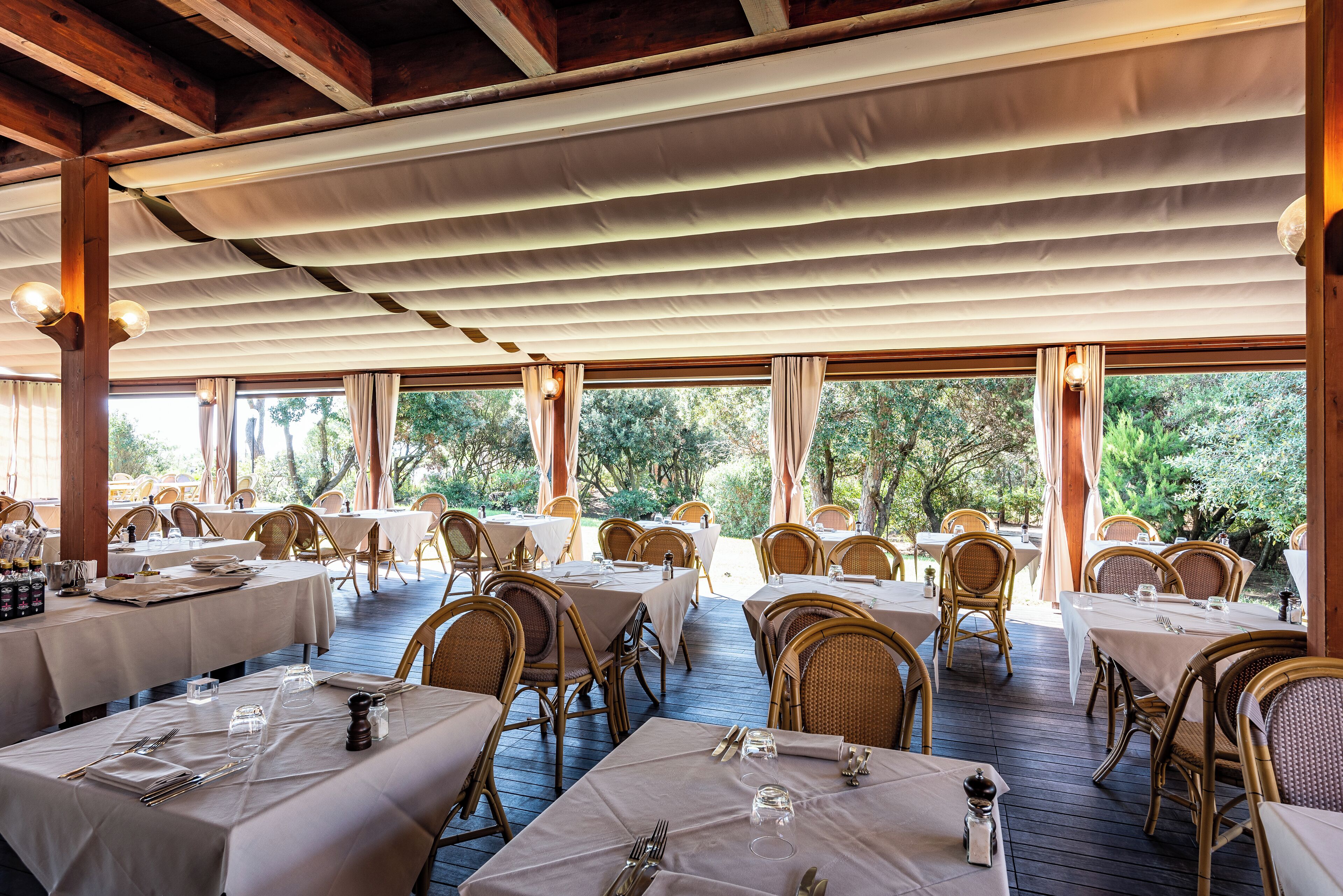 Un área de comedor al aire libre con mesas dispuestas para dos, rodeada de sillas de mimbre, bajo un techo cubierto con ventanas grandes.