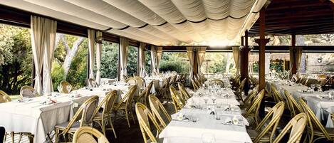 A restaurant with long tables, wicker chairs, and a covered outdoor area with curtains.