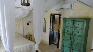A bedroom with a bed under a canopy, a green cabinet, an air conditioning unit, and a small bedside table with a lamp. - Mas du Puits d'Amour (Orange)