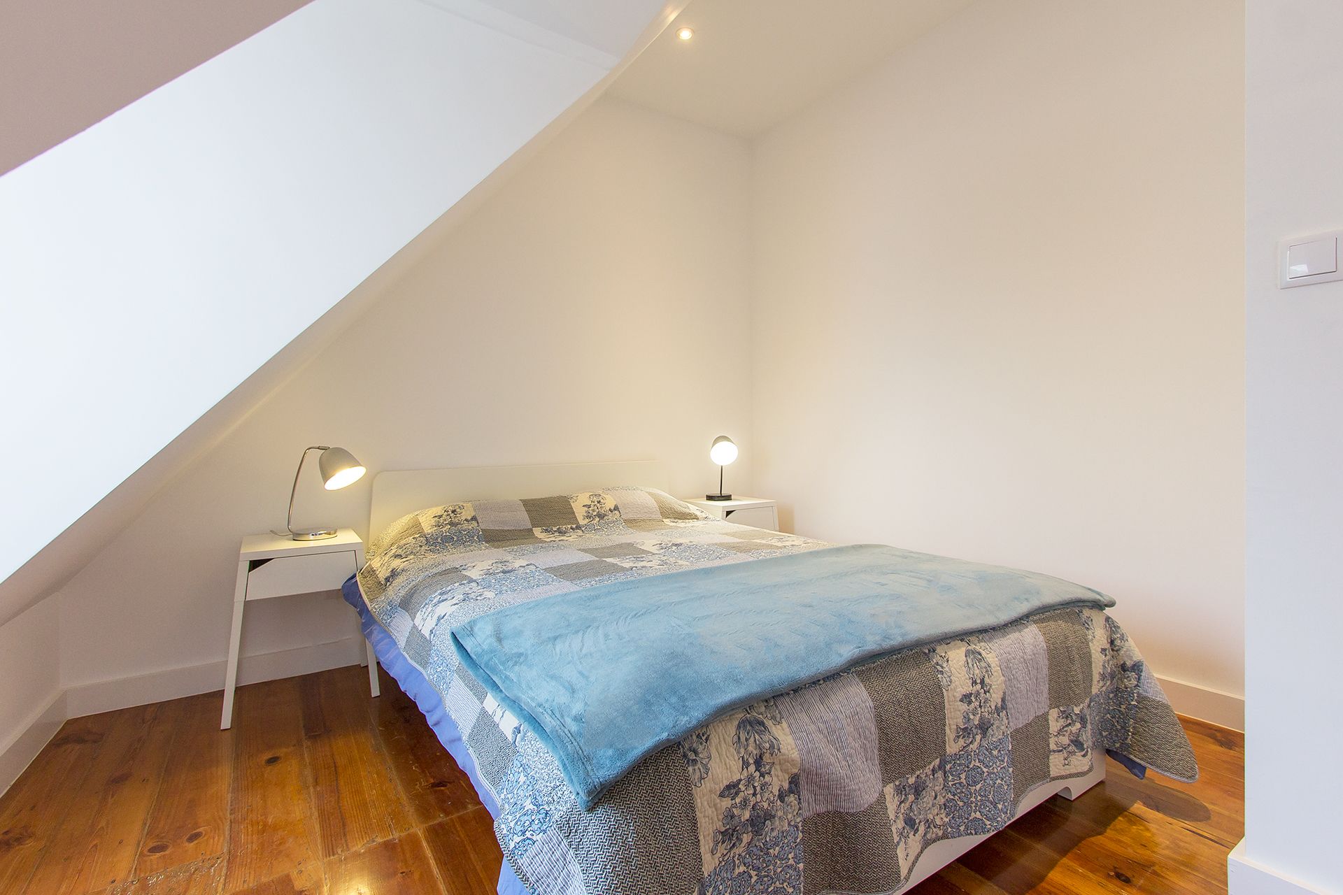 A bedroom with a sloped ceiling, a bed with patterned bedding, two bedside lamps, and wooden flooring.